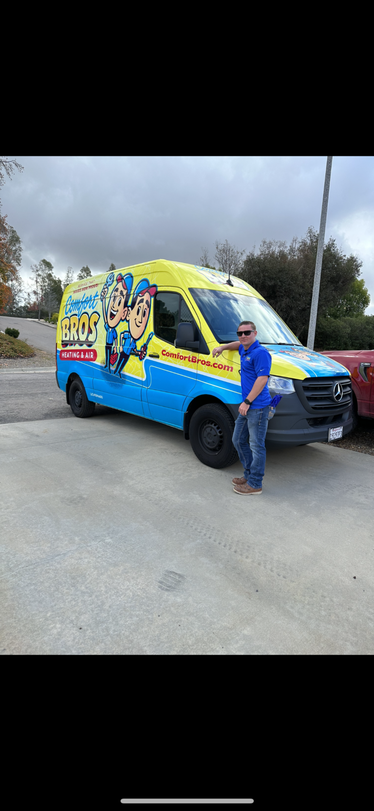 A man in sunglasses stands next to a blue and yellow Comfort Bros heating and air van parked on a concrete driveway.