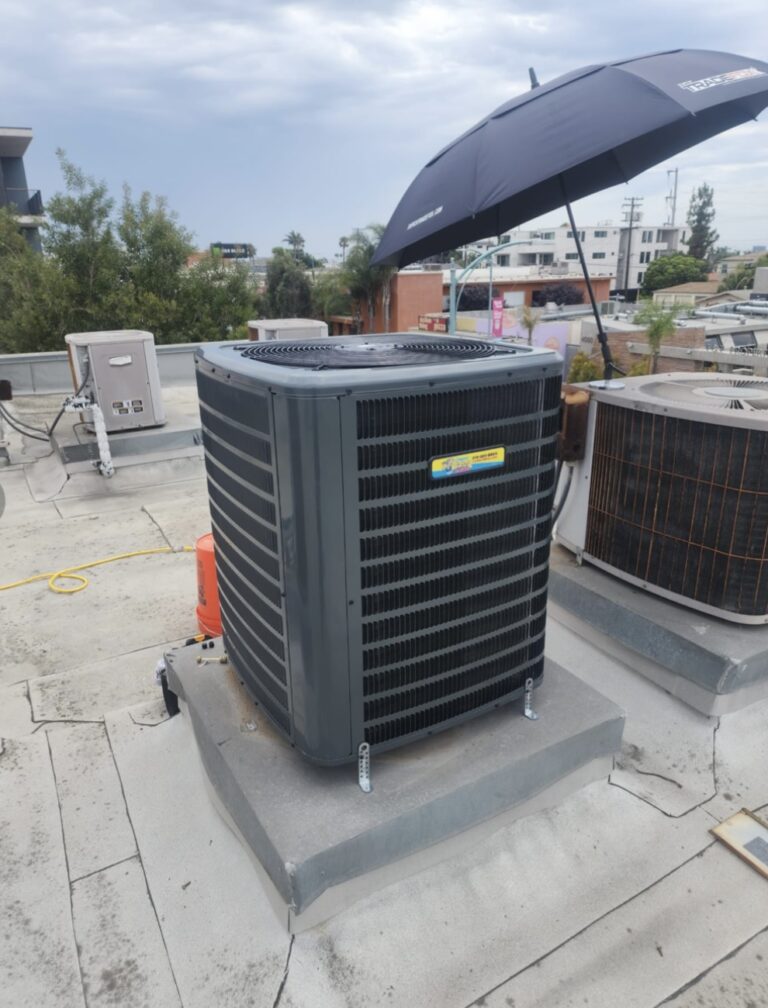 Large air conditioning unit on a rooftop, with a black umbrella providing shade overhead. Other HVAC equipment and buildings are visible in the background under a cloudy sky.
