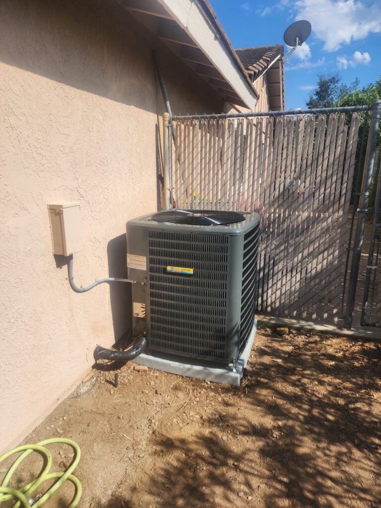 Outdoor air conditioning unit installed beside a house, next to a utility box and green garden hose, with a metal gate and fence in the background.