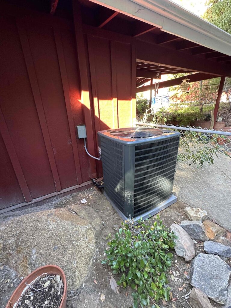 A black outdoor air conditioning unit sits on a concrete pad next to a red wooden house, near a chain-link fence and some plants.