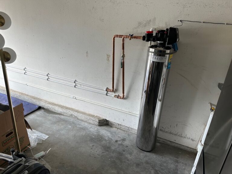A water filtration system with copper pipes is installed on a garage wall, with pipes running horizontally and a cardboard box on the floor nearby.