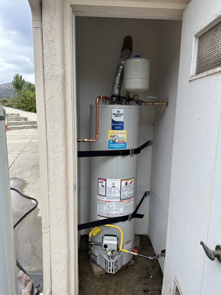 A water heater is installed in a small outdoor closet with visible pipes, warning labels, and a black strap securing the tank.