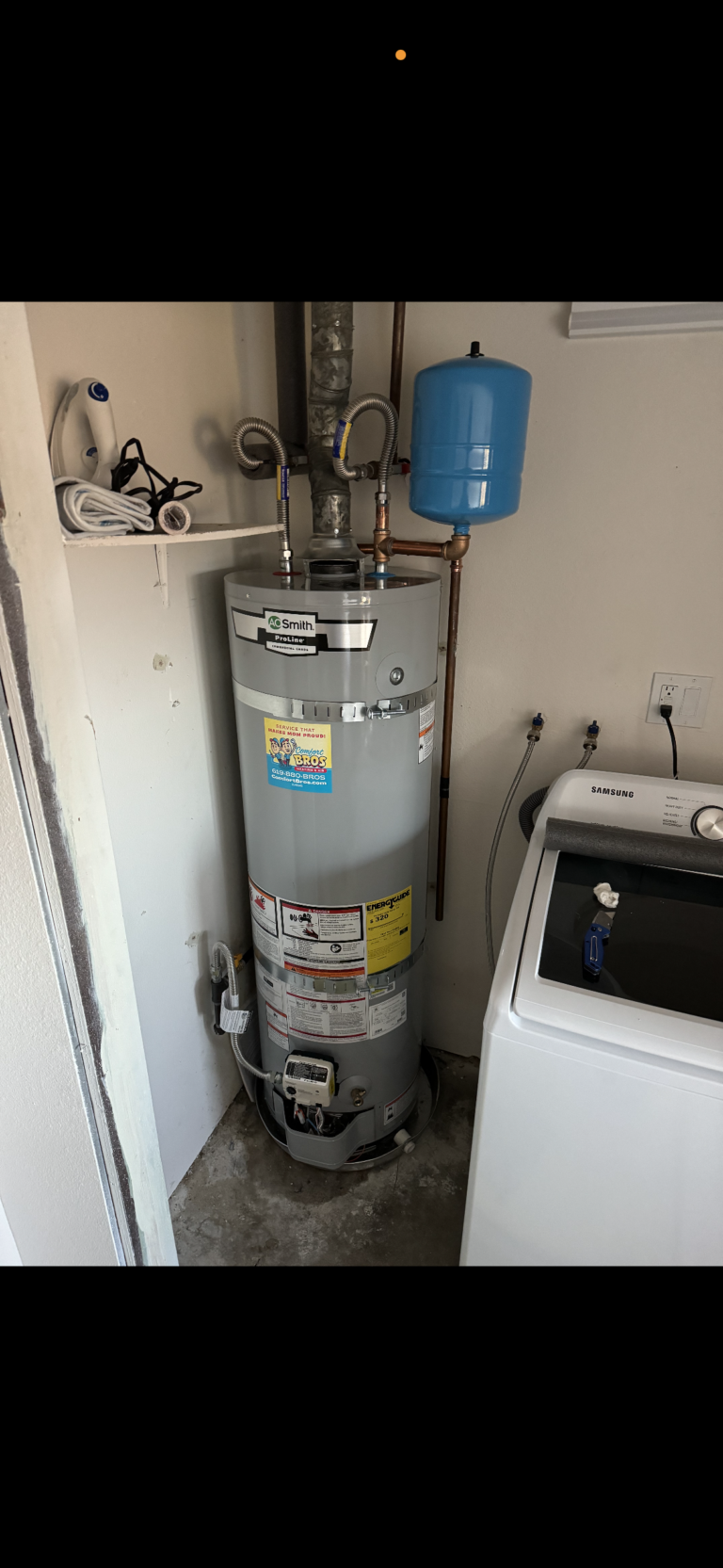 A water heater with attached pipes and a blue expansion tank is installed next to a washing machine in a laundry room.
