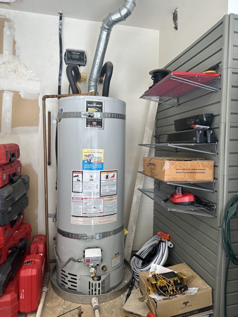 A gray water heater is installed in a utility room, surrounded by red toolboxes, extension cords, shelves, and various tools and equipment.