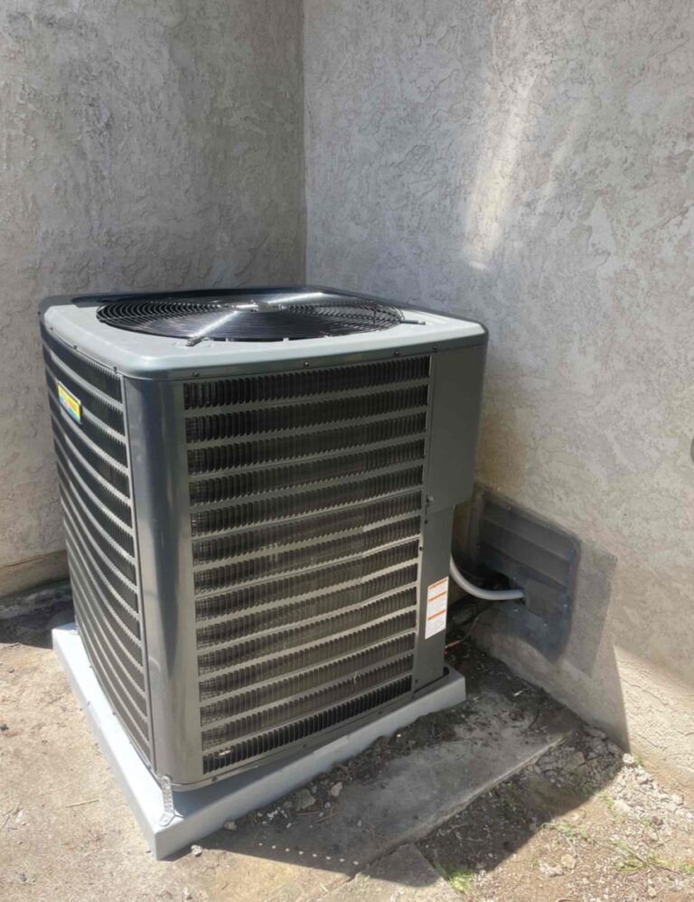 Outdoor air conditioning unit placed on a concrete pad next to a stucco wall, with electrical and coolant lines connected through the wall.