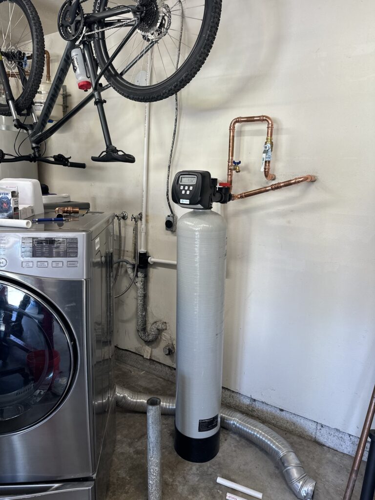 A water softener system with copper pipes is installed next to a washing machine and a bicycle in a garage.