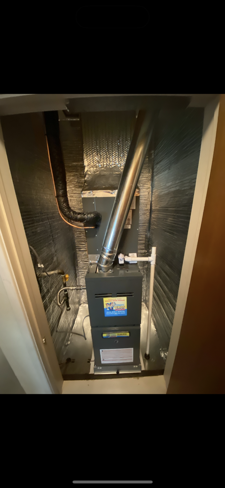 A residential furnace with attached ductwork and pipes is installed in a small utility closet with metallic insulated walls.