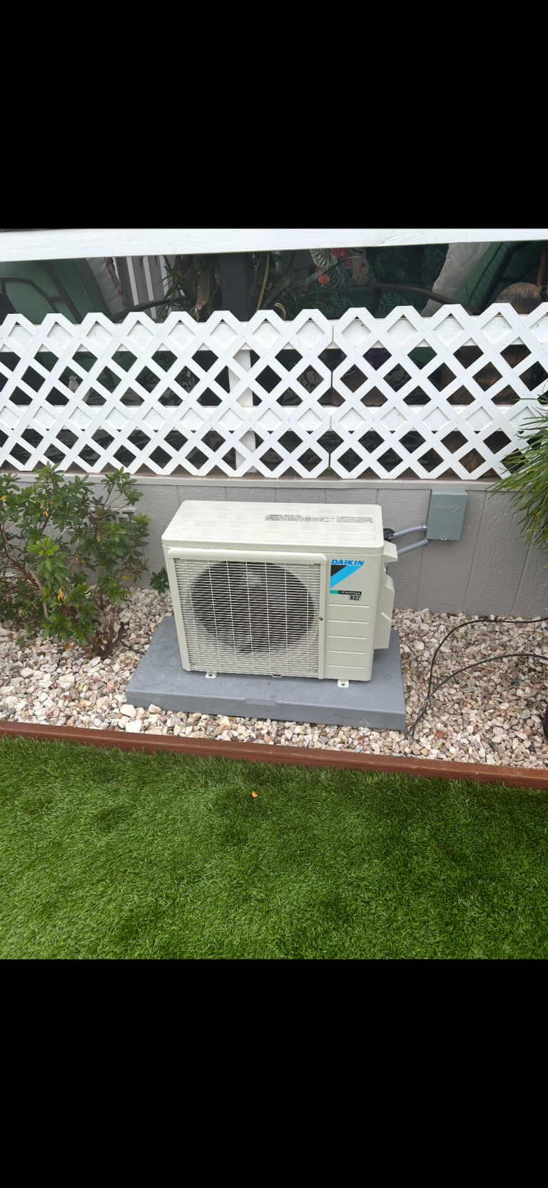 Outdoor air conditioning unit installed on a concrete pad, surrounded by gravel, plants, and artificial grass, with a white lattice fence in the background.