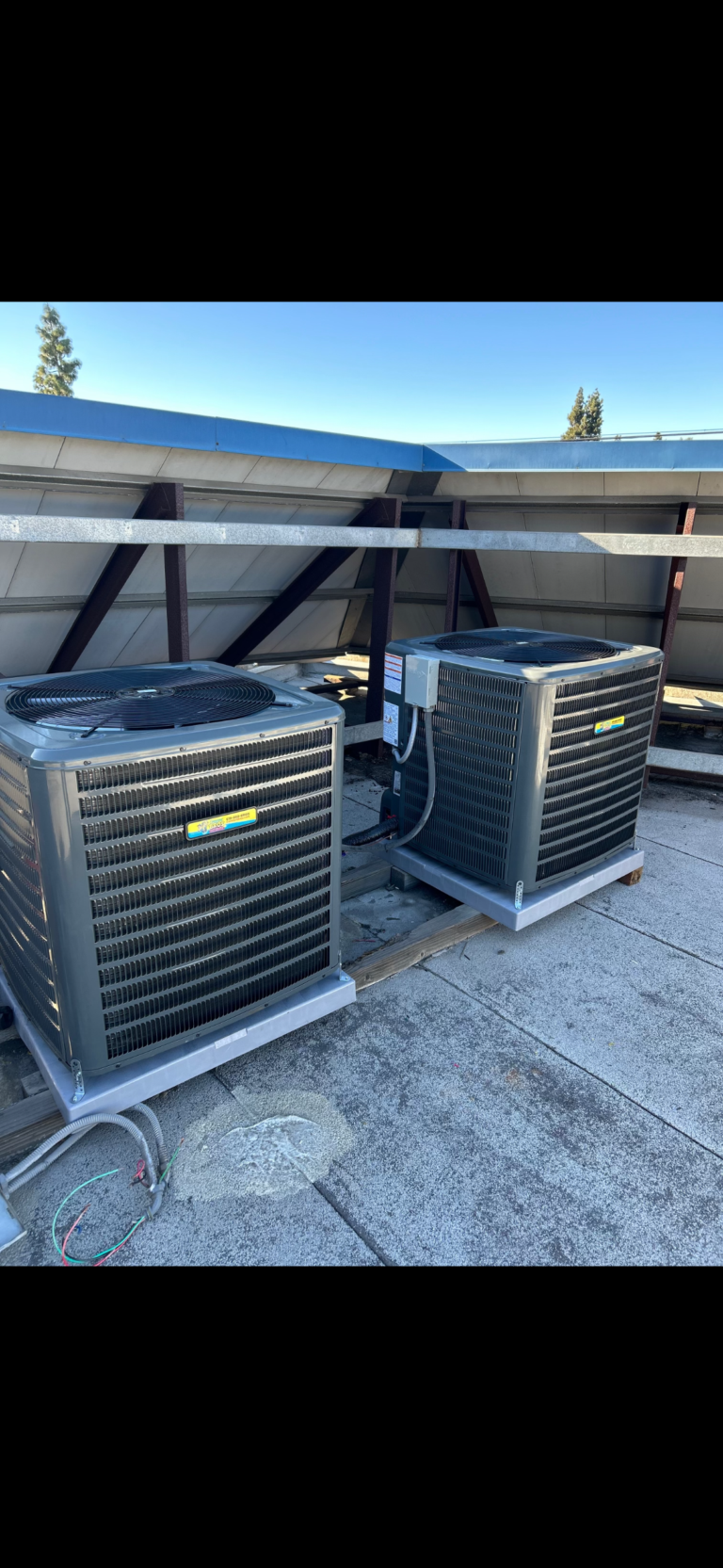 Two large outdoor air conditioning units are installed on a rooftop, connected by cables and sitting on a concrete surface under a clear sky.