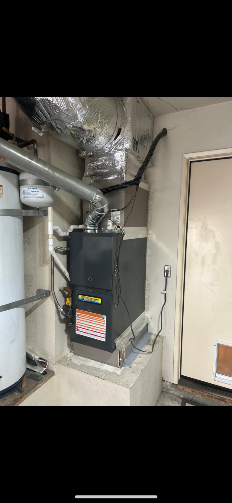 A furnace with attached ductwork and nearby water heater is installed on a raised platform in a utility room next to a door with a pet door at the bottom.