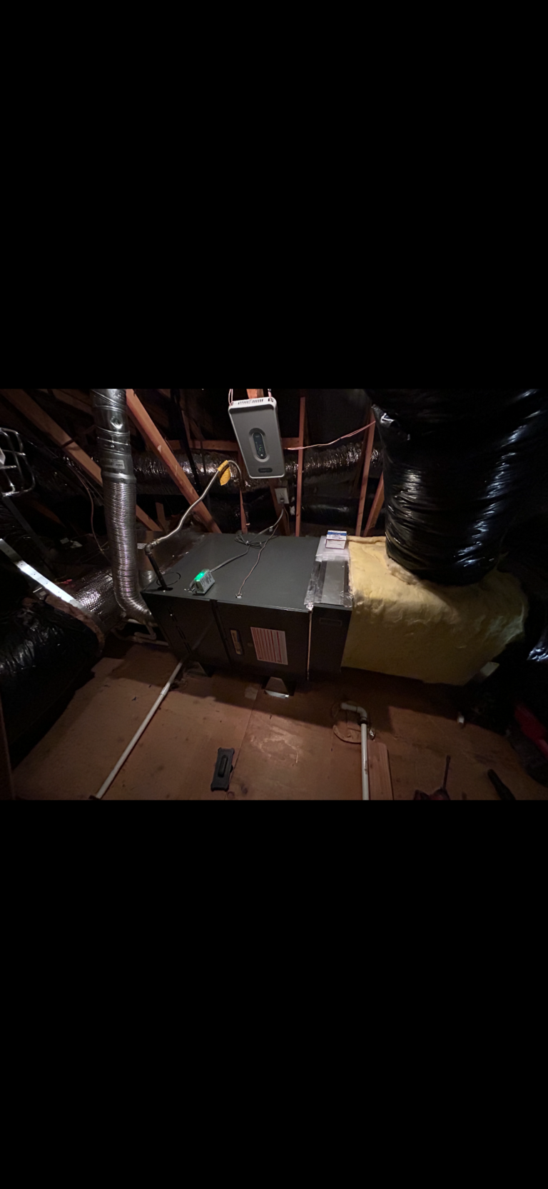 A heating and air conditioning unit is installed in an attic with exposed insulation, ducts, pipes, and electrical wiring visible around the unit.