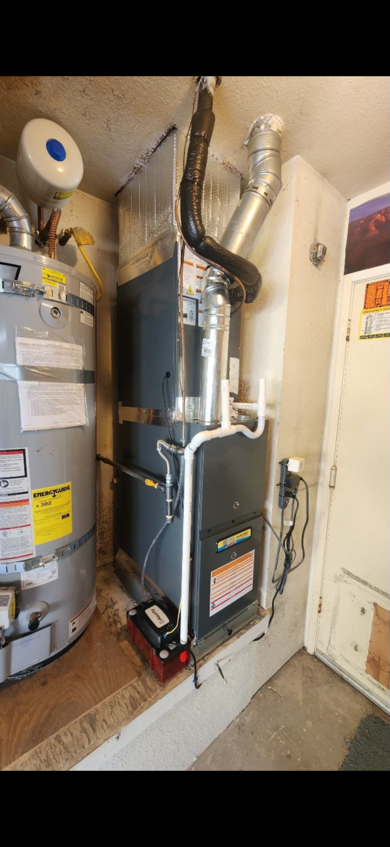 A water heater and furnace unit installed side by side in a garage, with visible pipes, ducts, labels, and a battery-powered device on the floor.
