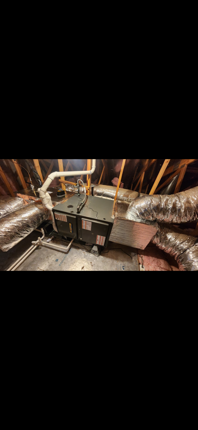 Residential HVAC unit installed in an attic, surrounded by insulated air ducts and exposed wooden beams.