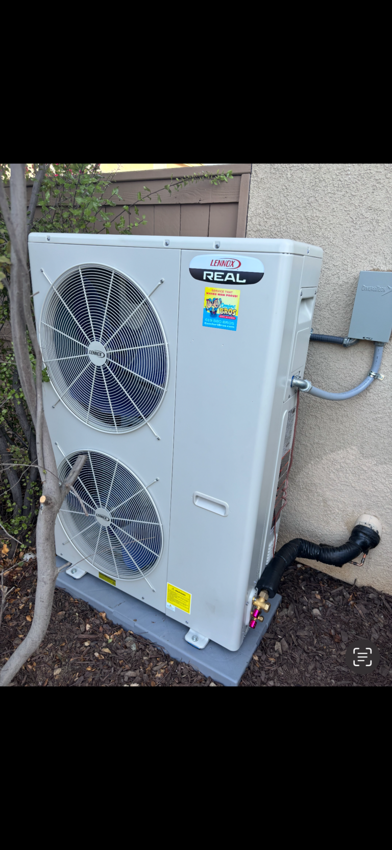 Outdoor air conditioning unit with two large fans installed on a concrete pad next to a stucco wall, with electrical and refrigerant lines connected.