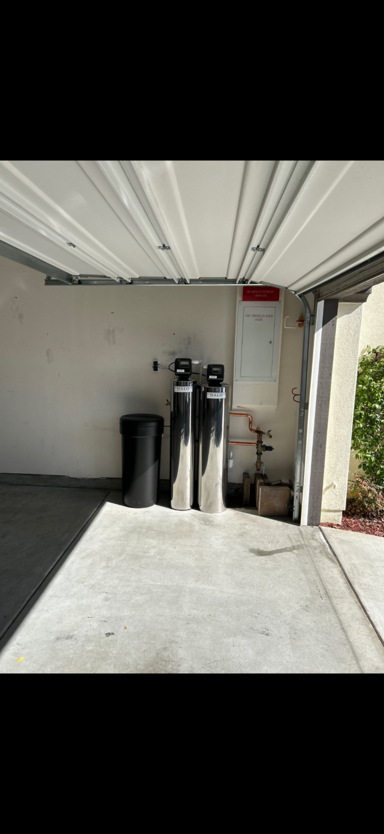 Open garage with two water softener tanks, a black trash bin, a white control panel on the wall, and sunlight casting shadows on the concrete floor.