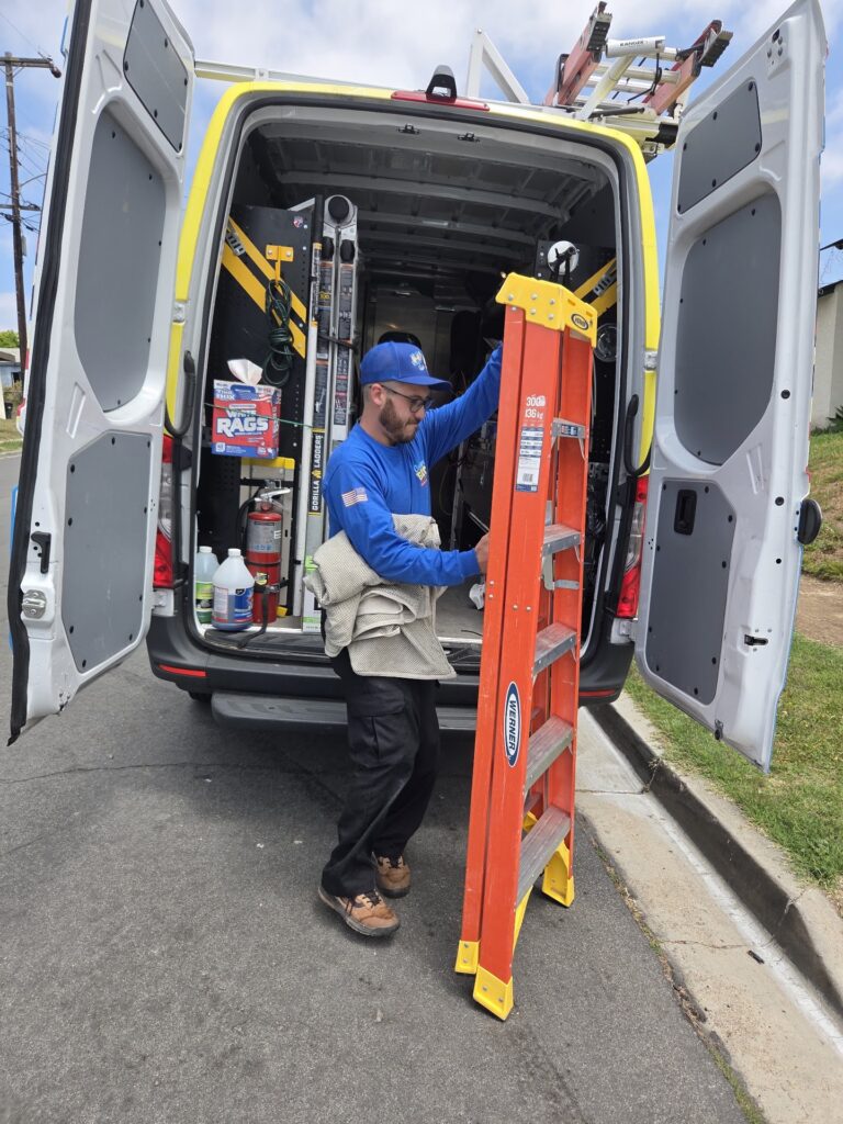 A worker in a blue shirt and cap unloads an orange ladder from the back of a work van filled with tools and equipment on a residential street.