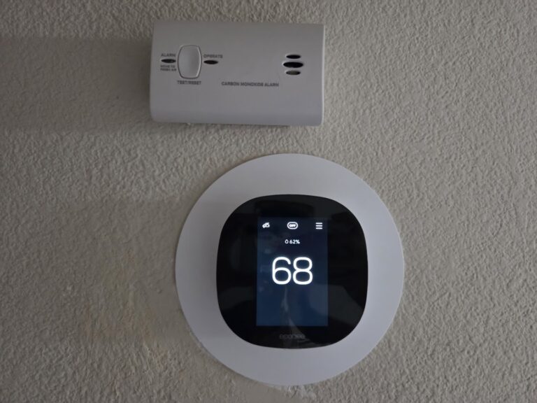 Wall-mounted carbon monoxide alarm above a digital thermostat showing 68 degrees, both attached to a beige textured wall.