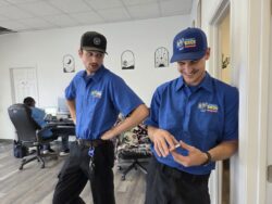 Two men in blue uniforms stand and smile in an office, while another person works at a computer in the background.