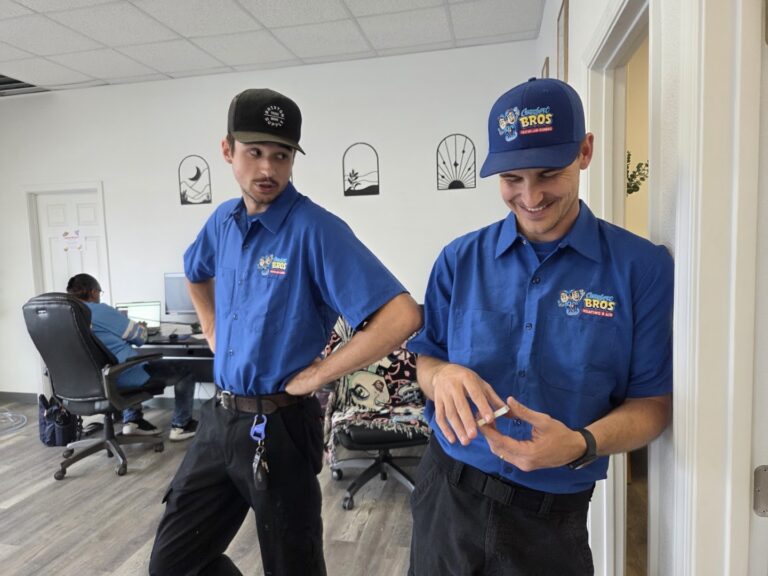 Two men in blue uniforms stand and smile in an office, while another person works at a computer in the background.