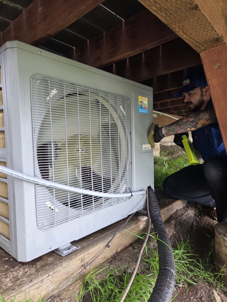 A person wearing gloves cleans an outdoor HVAC unit under a deck using a cloth and spray bottle.