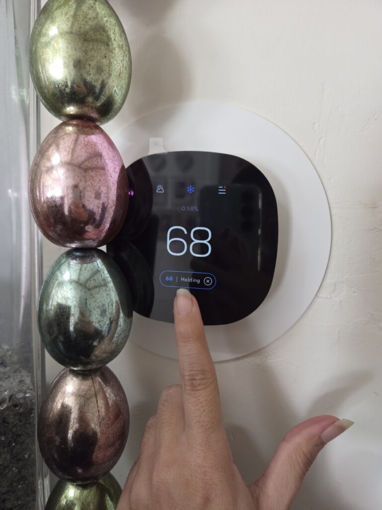 A hand with long nails sets a digital thermostat to 68 degrees Fahrenheit. Metallic decorative objects are visible on the left side of the image.