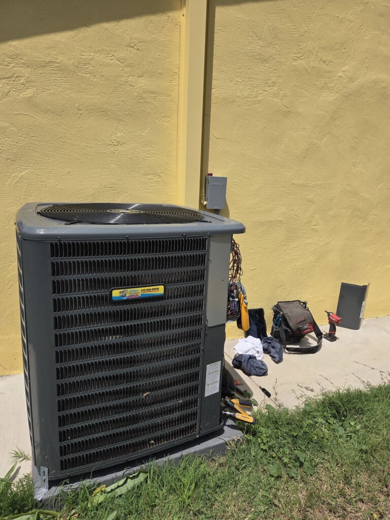 A large air conditioning unit is installed beside a yellow wall, with various tools, a tool bag, and a removed panel placed on the ground nearby.