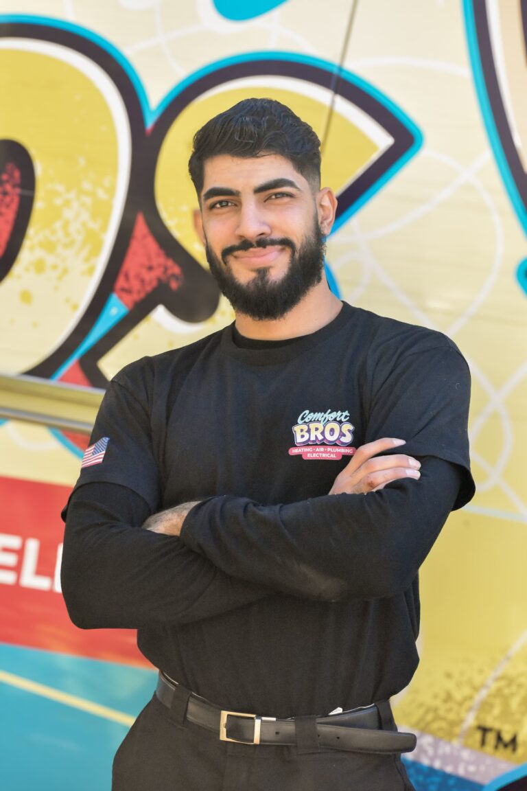 A man with dark hair and a beard stands in front of a colorful mural, wearing a “Comfort Bros” shirt and smiling with his arms crossed.