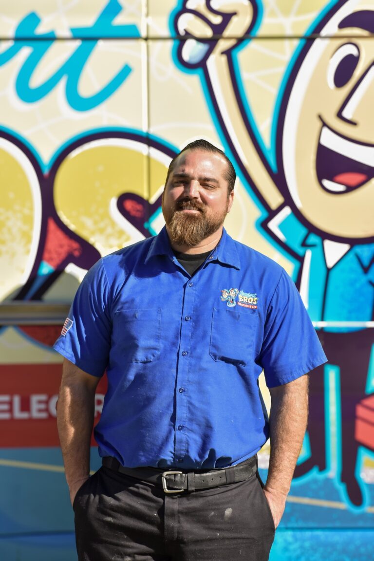 A man in a blue button-up shirt stands in front of a colorful vehicle with cartoon graphics and text, looking at the camera with hands in his pockets.