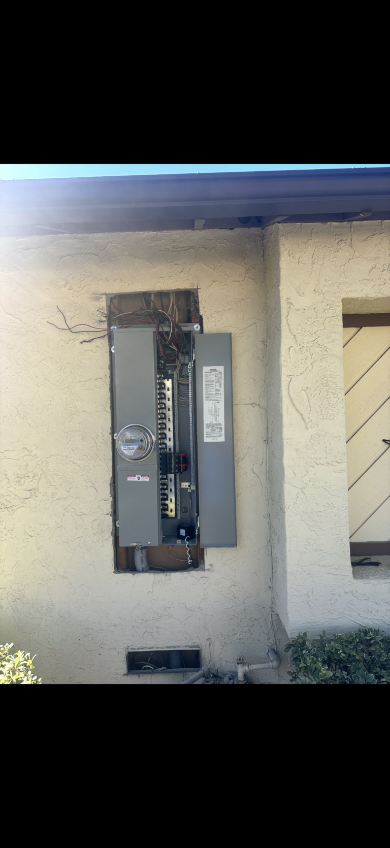 Outdoor electrical panel with the cover open on a stucco wall, exposing circuit breakers and wiring inside.