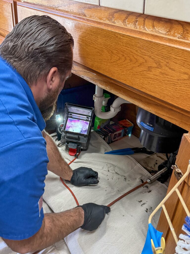 A plumber uses a camera and inspection tool to examine pipes under a kitchen sink with cleaning supplies and a white mat on the floor.