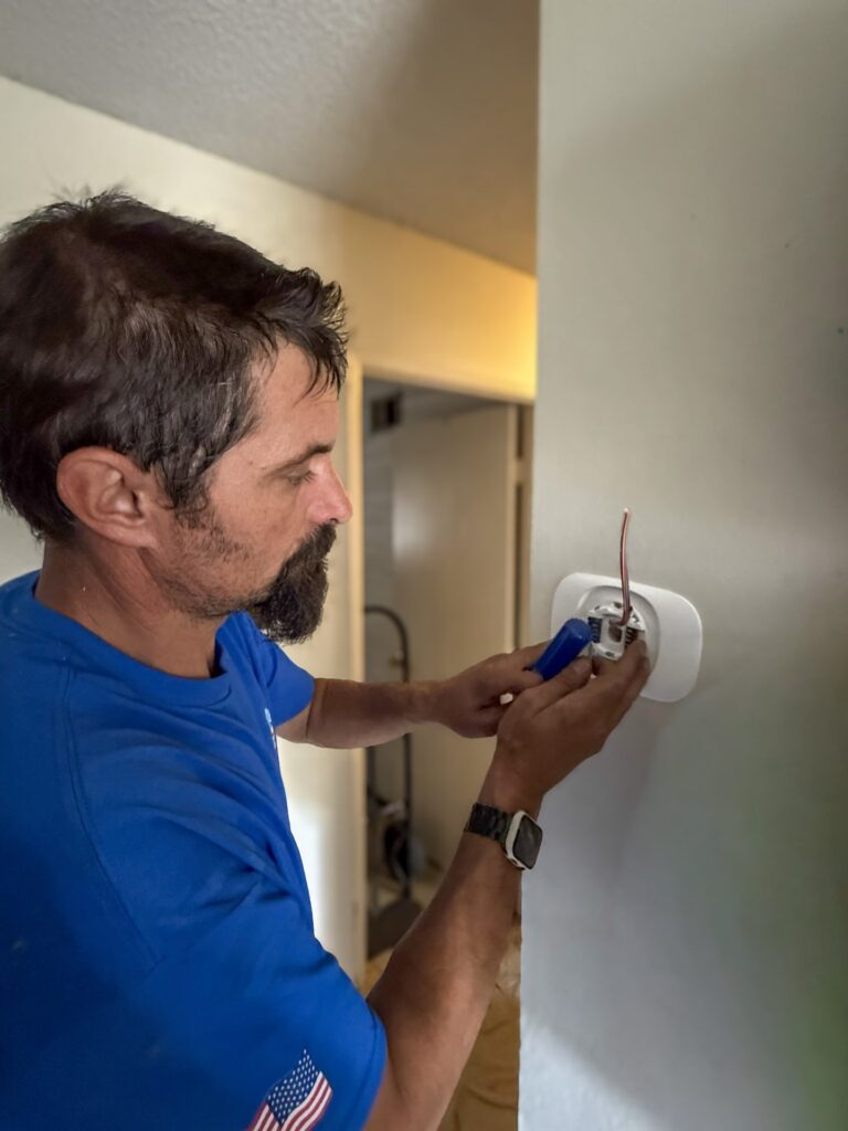 Man in a blue shirt installs a thermostat on a white wall, using a screwdriver to connect wires.