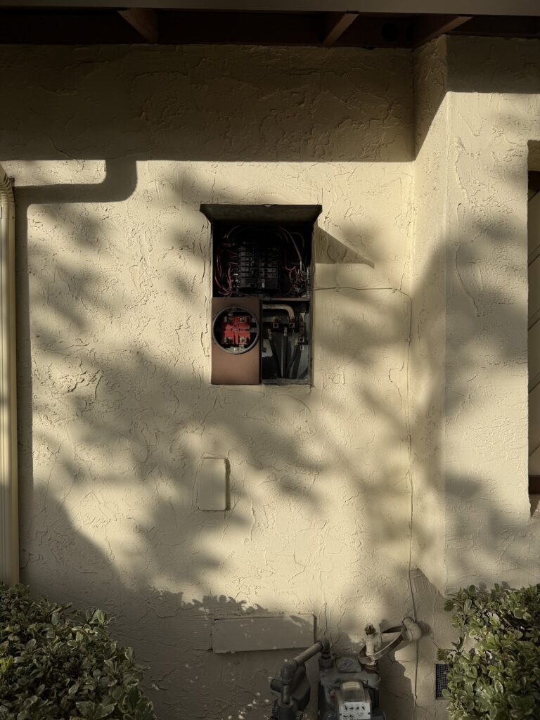 An electrical panel with its cover open is mounted on a stucco exterior wall of a building, exposing wiring and components inside. Bushes line the bottom of the wall.