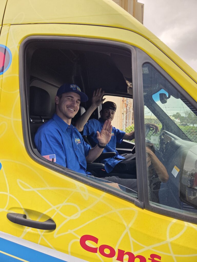 Two people in blue uniforms sit in the front seats of a yellow van, smiling and waving at the camera through the open driver’s side window.