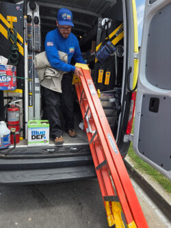 A worker in a blue shirt and hat loads an orange ladder into the back of a service van equipped with tools and supplies.