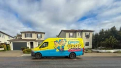A brightly colored Comfort Bros. van is parked on a suburban street in front of two modern houses under a cloudy sky.