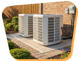 Two outdoor air conditioning units sit on a concrete pad next to a brick house, surrounded by landscaping and a wooden fence.