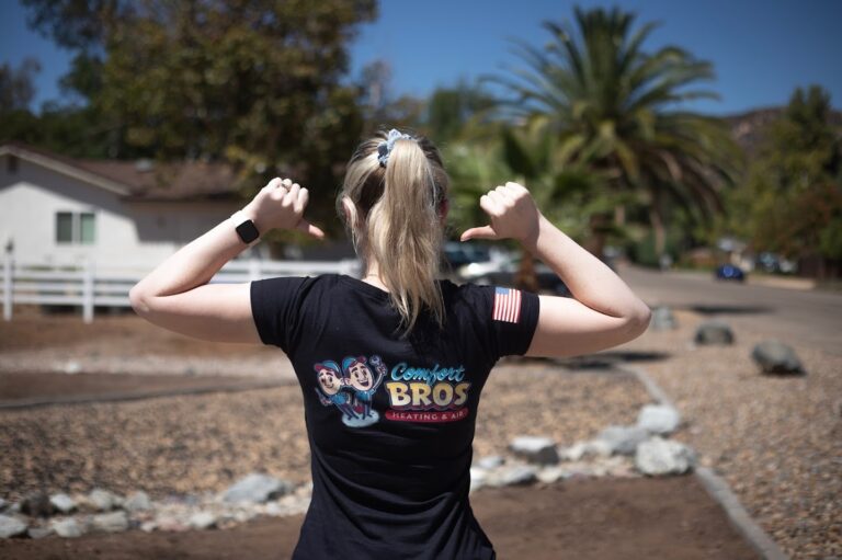 A person with a ponytail points to the logo on the back of their “Comfort Bros Heating & Air” shirt while standing on a sunny suburban street.