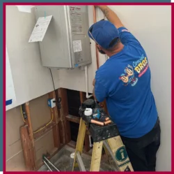 A plumber in a blue shirt installs or repairs a wall-mounted tankless water heater in a partially finished room, standing on a step ladder.