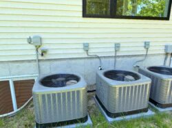 Three outdoor air conditioning units are installed next to the exterior wall of a house beneath a window.