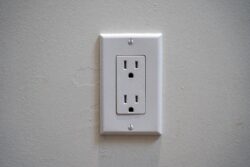 A standard white electrical wall outlet with two sockets is mounted on a plain light-colored wall.