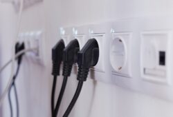 Several black electrical plugs are inserted into a row of white wall outlets, with a few unused sockets visible.
