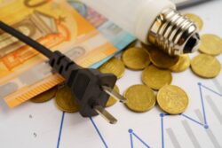 A power plug, light bulb, coins, and banknotes are arranged on a financial chart with blue lines.