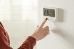A person adjusts a digital wall thermostat, displaying a temperature of 21°C and the time 23:15, using the buttons below the screen.